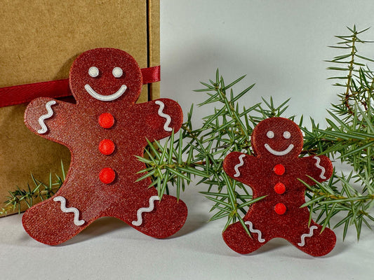 Two red gingerbread man ornaments on a white surface with a gift and greenery in the background.