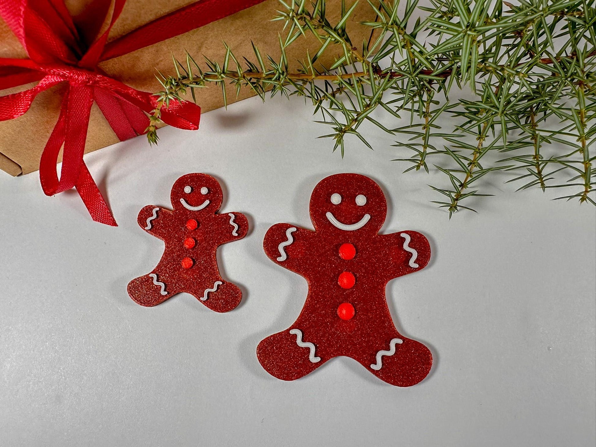 Two red gingerbread man ornaments on a white surface with a gift and greenery in the background.