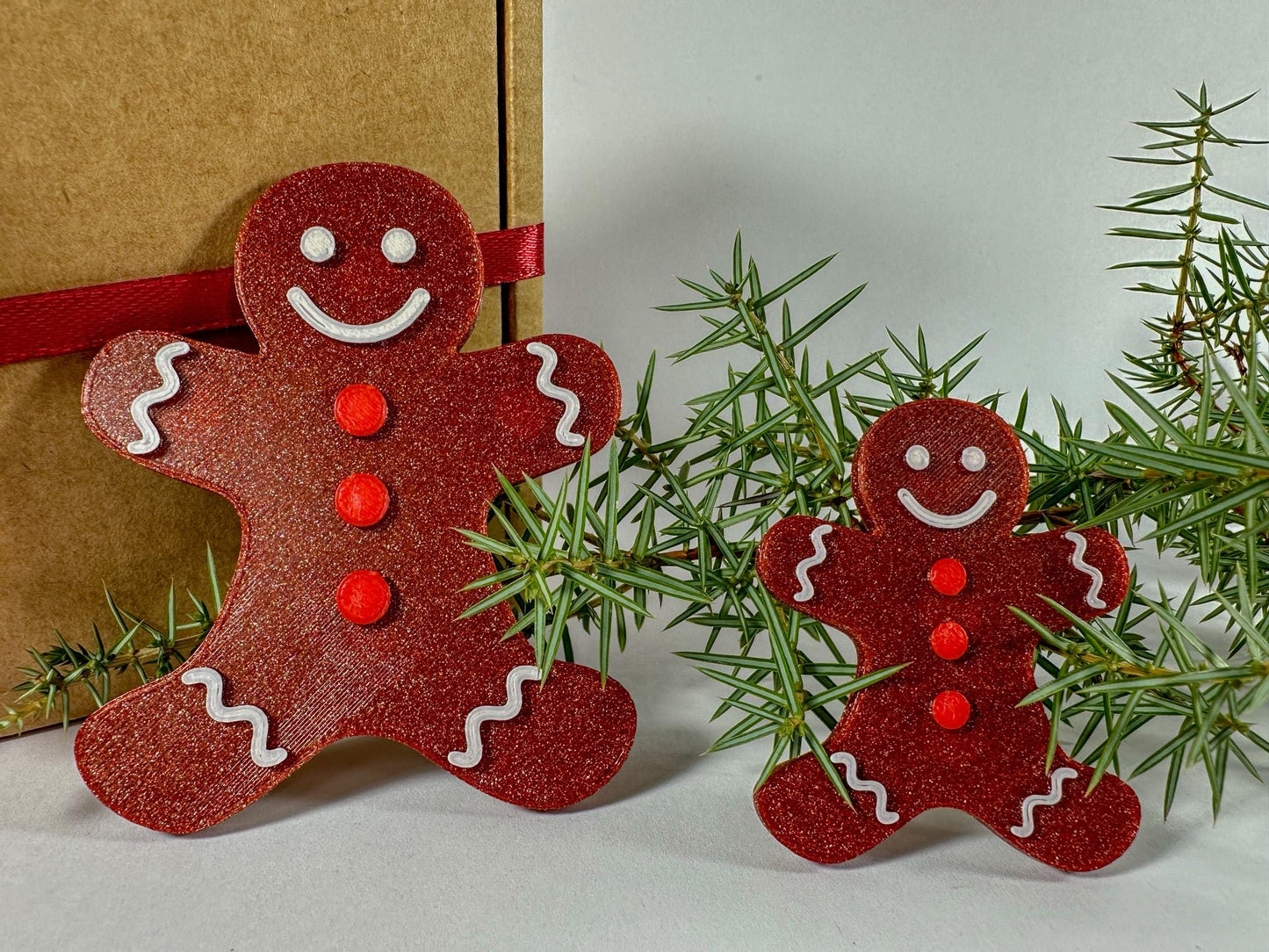 Two red gingerbread man ornaments on a white surface with a gift and greenery in the background.