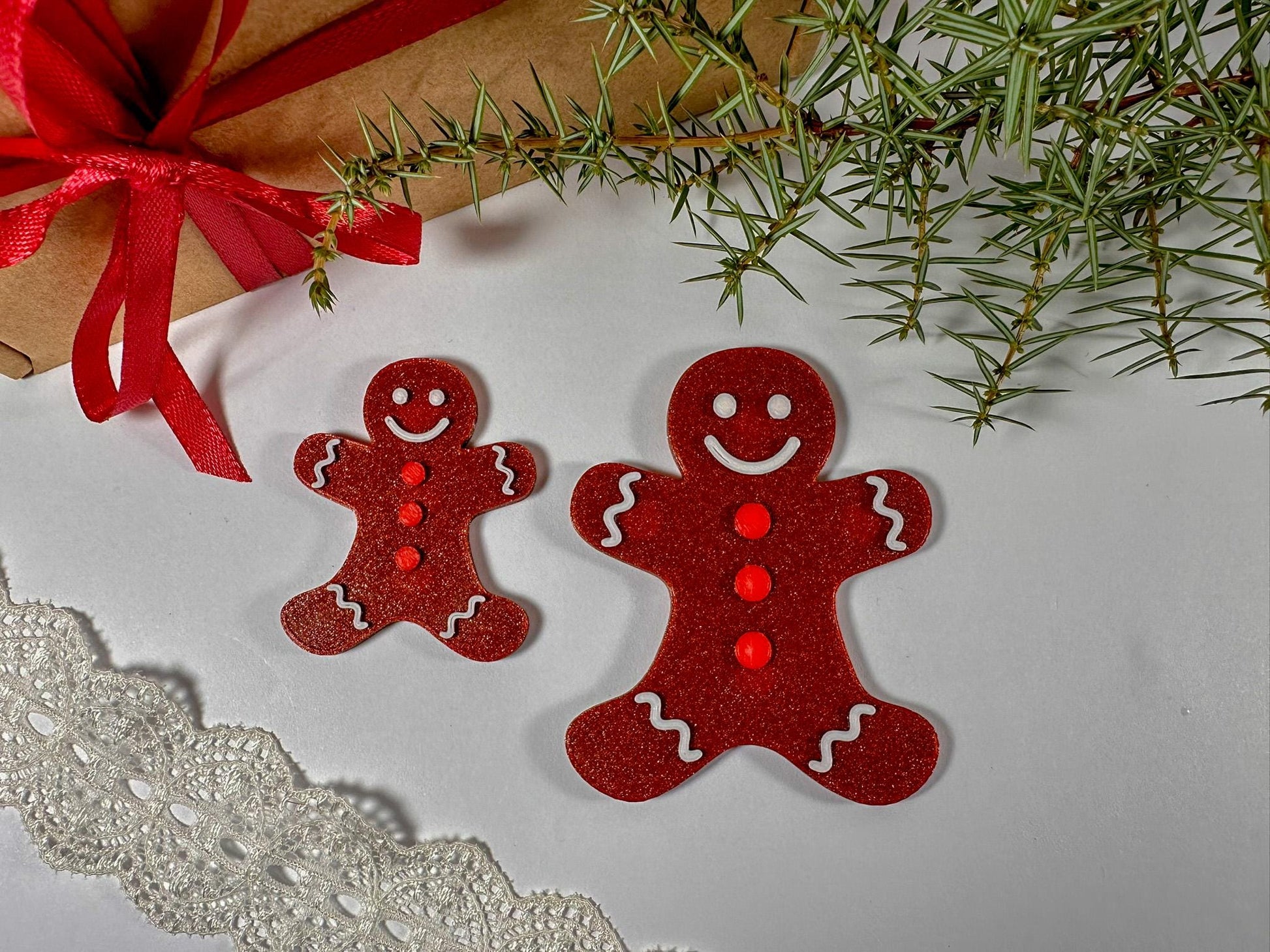 Two red gingerbread man ornaments on a white surface with a gift and greenery in the background.