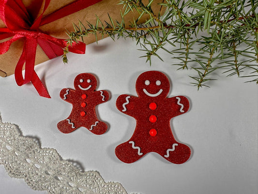 Two red gingerbread man ornaments on a white surface with a gift and greenery in the background.